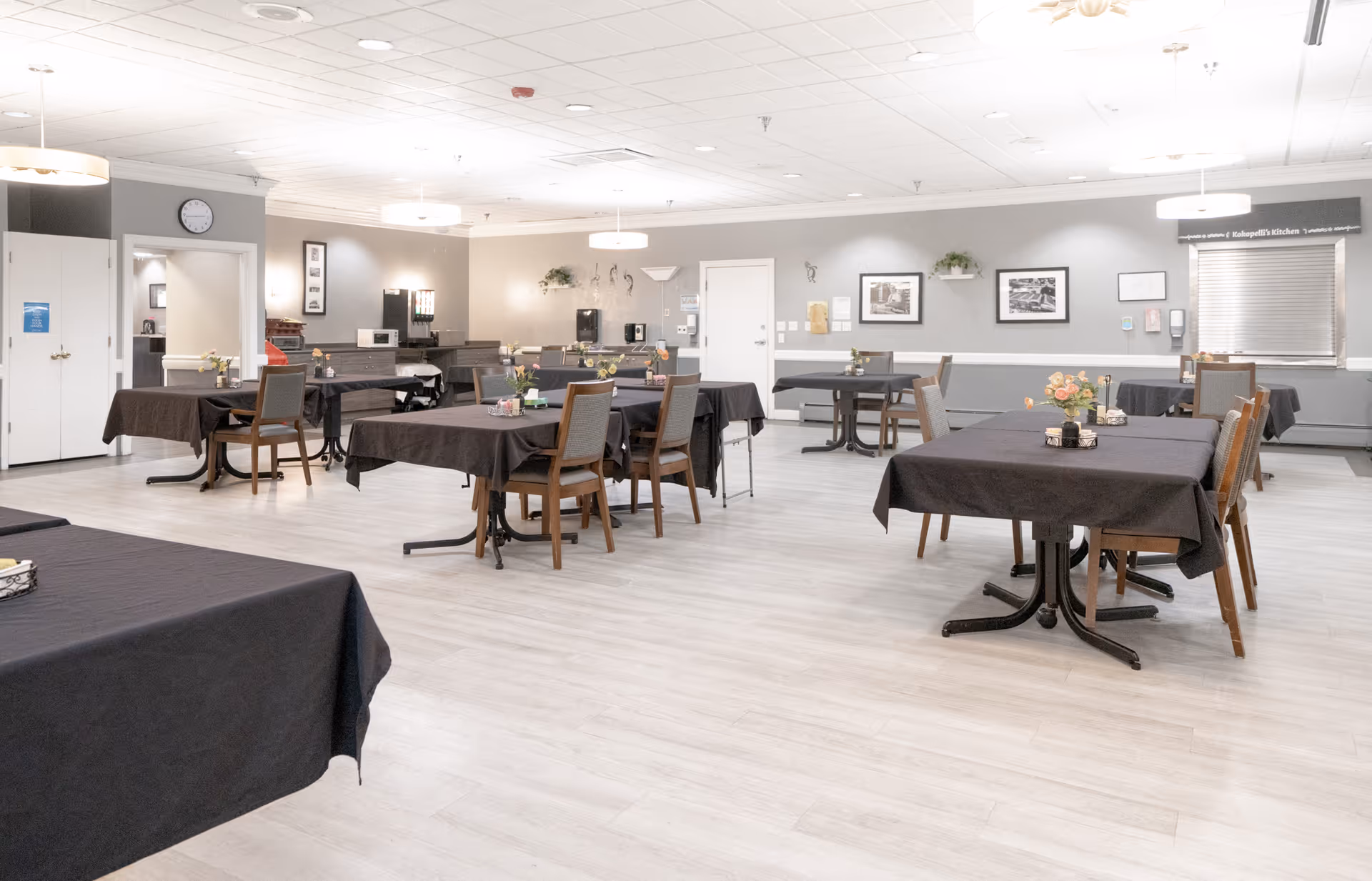 A spacious dining room with multiple tables covered in dark tablecloths, each surrounded by wooden chairs. The room has light-colored flooring, neutral walls with framed pictures, and ceiling lights. There is a service window on the right side and a clock on the wall near a doorway on the left.