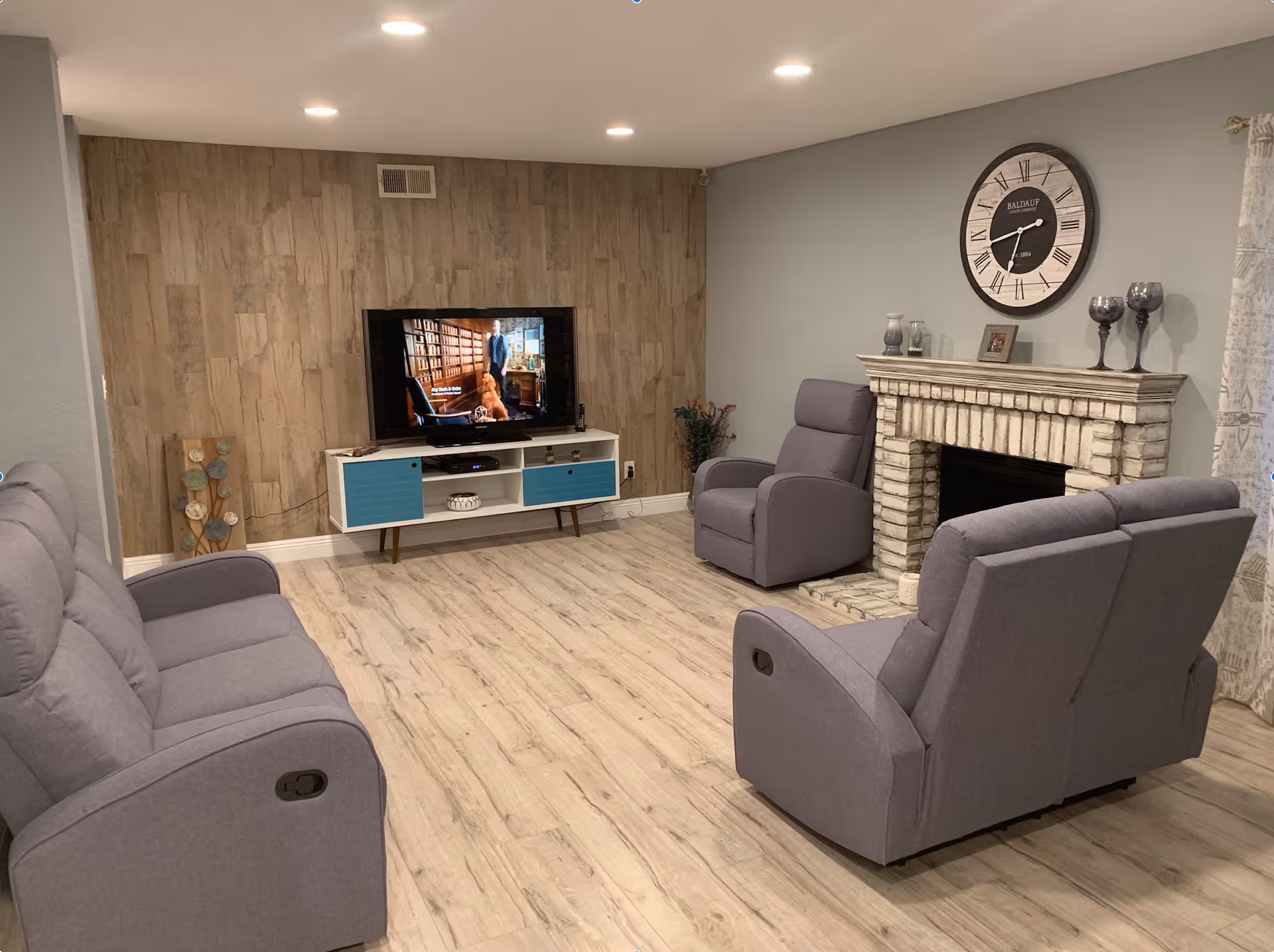 A cozy living room with light wood flooring, a wooden accent wall behind a flat-screen TV on a white and blue TV stand, three gray recliner chairs arranged around a white brick fireplace with decorative items on the mantel, and a large round wall clock above the fireplace.