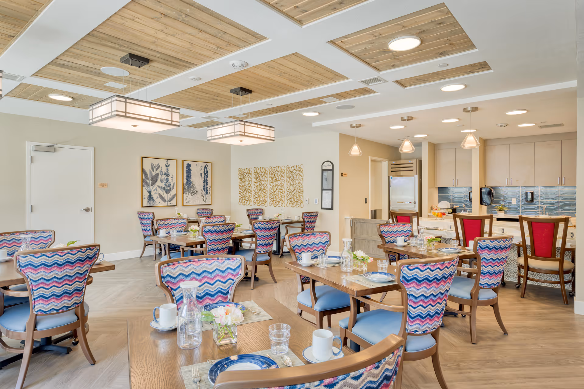 Bright and modern dining room with wooden tables set with plates, cups, and glassware. Colorful chairs with zigzag patterned upholstery and blue seats surround the tables. The ceiling features wooden panels with recessed lighting and two large rectangular pendant lights. The back wall has framed botanical artwork and decorative wall panels. A kitchen area with cabinets, a refrigerator, and a blue tiled backsplash is visible in the background.