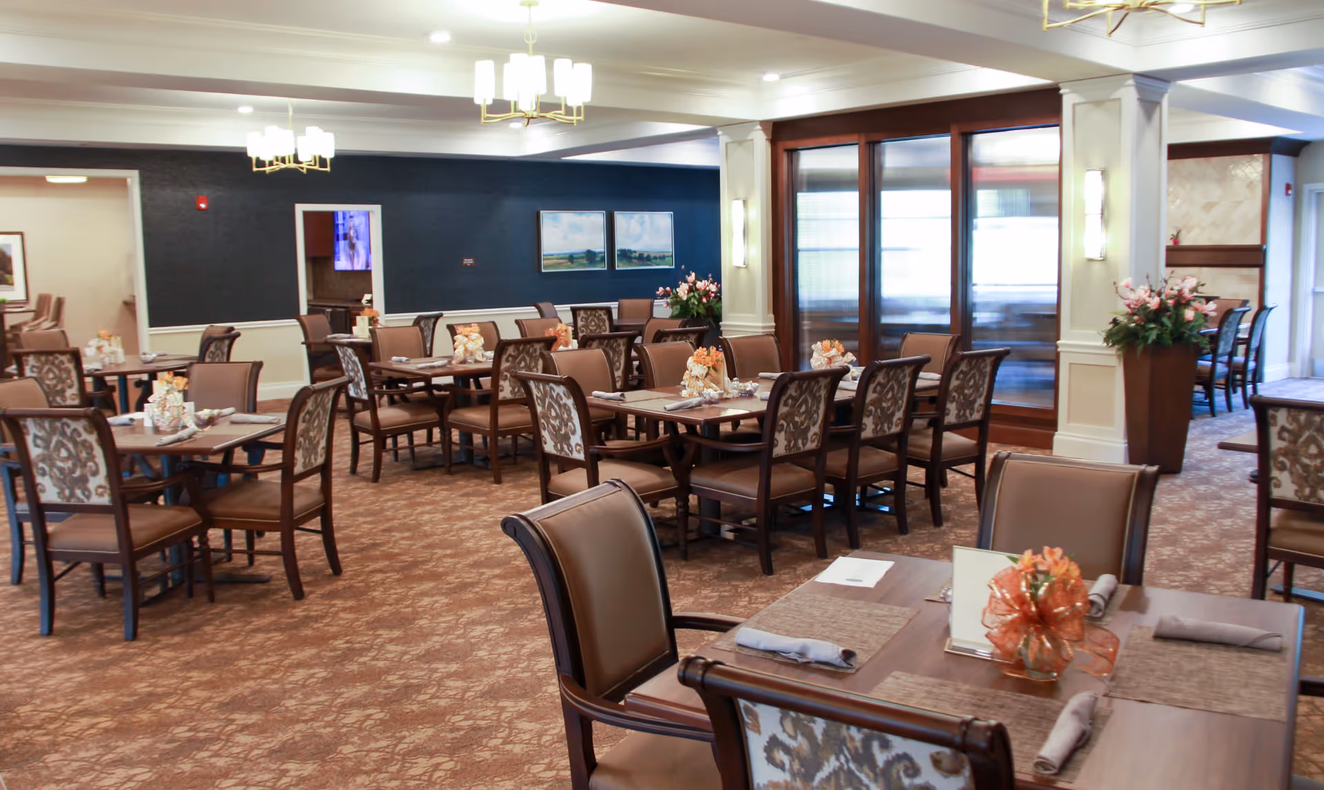 A spacious dining room in a senior living facility with multiple tables and chairs arranged neatly. Each table is set with placemats, napkins, and decorative centerpieces. The room features carpeted flooring, elegant light fixtures, and large windows with wooden frames allowing natural light to enter. There are paintings on the dark blue wall and floral arrangements placed around the room.