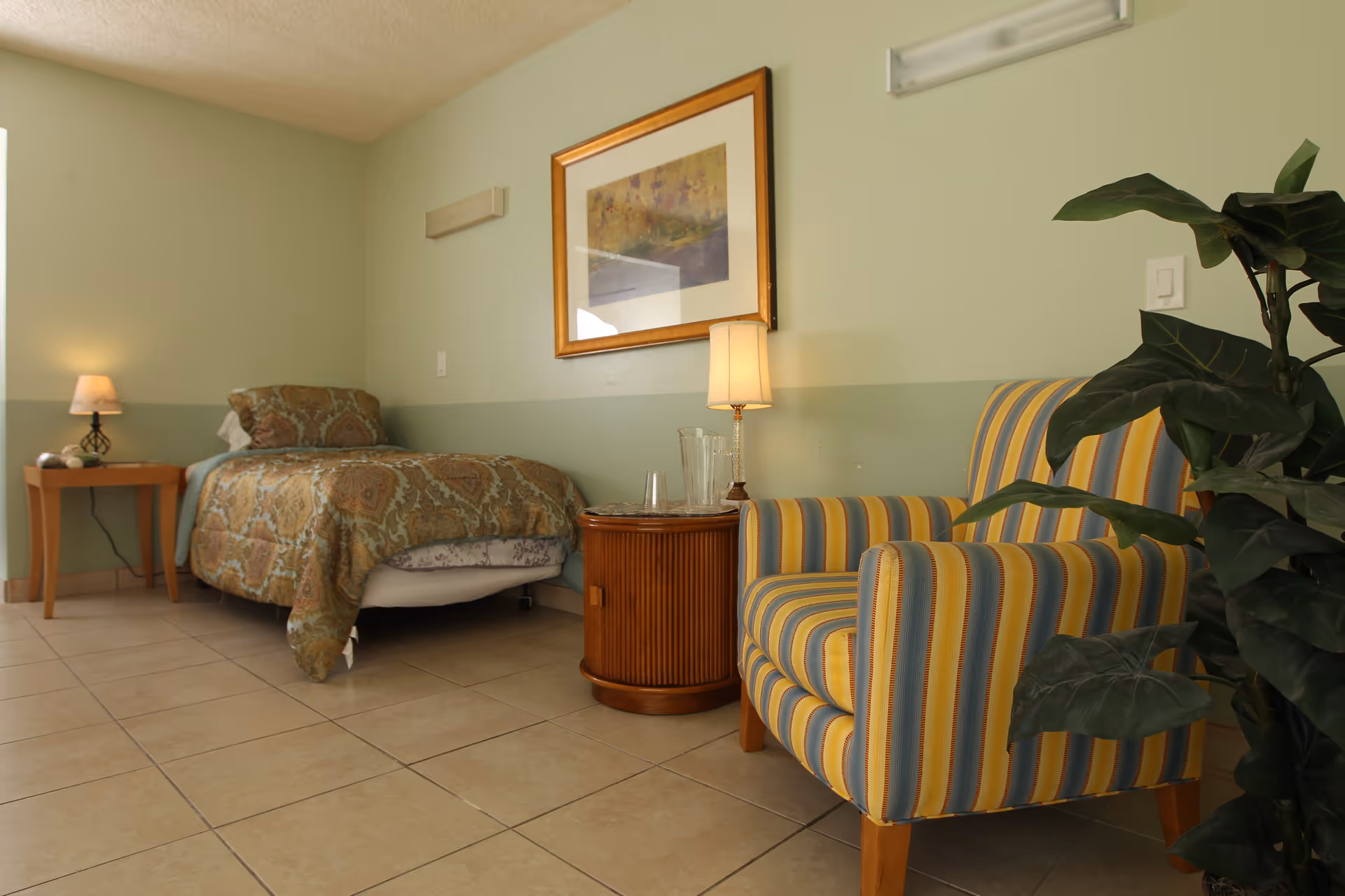 A furnished senior living bedroom with a single bed, bedside tables and lamp, a striped armchair, potted plant, and framed artwork on the wall.