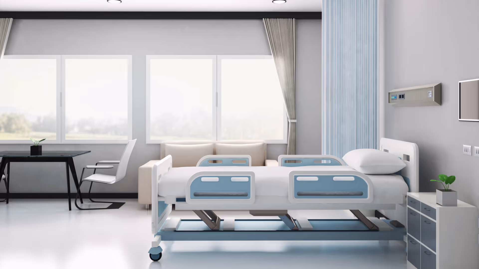 A bright modern patient room with a hospital bed, sofa, desk, and large windows.