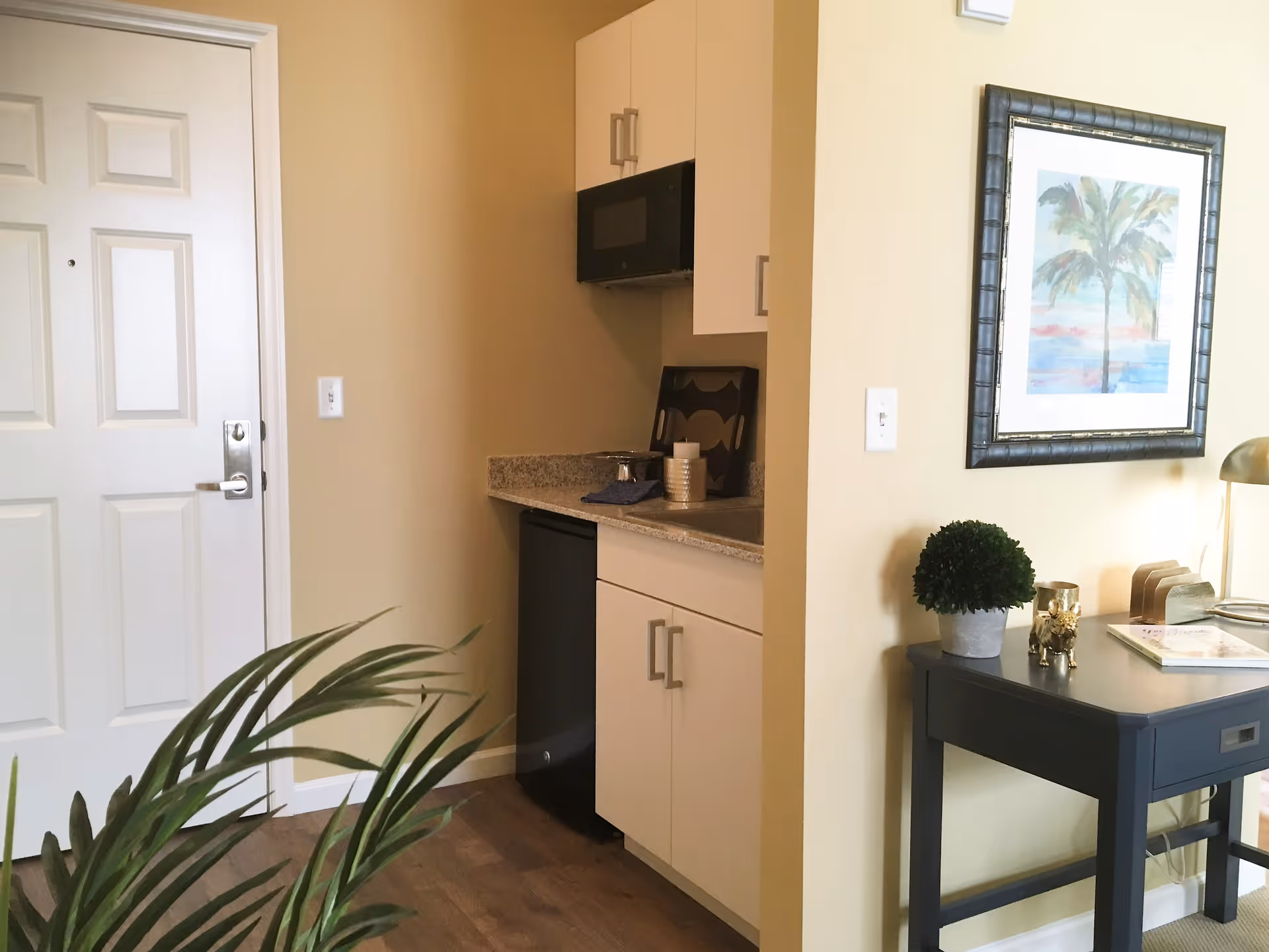 Small kitchenette area with white cabinets, a black microwave, and a countertop with a few items on it. To the left is a white door, and to the right is a dark gray desk with a small potted plant, a gold decorative item, a lamp, and some papers. A framed picture of a palm tree hangs on the wall above the desk. A green plant is partially visible in the foreground.