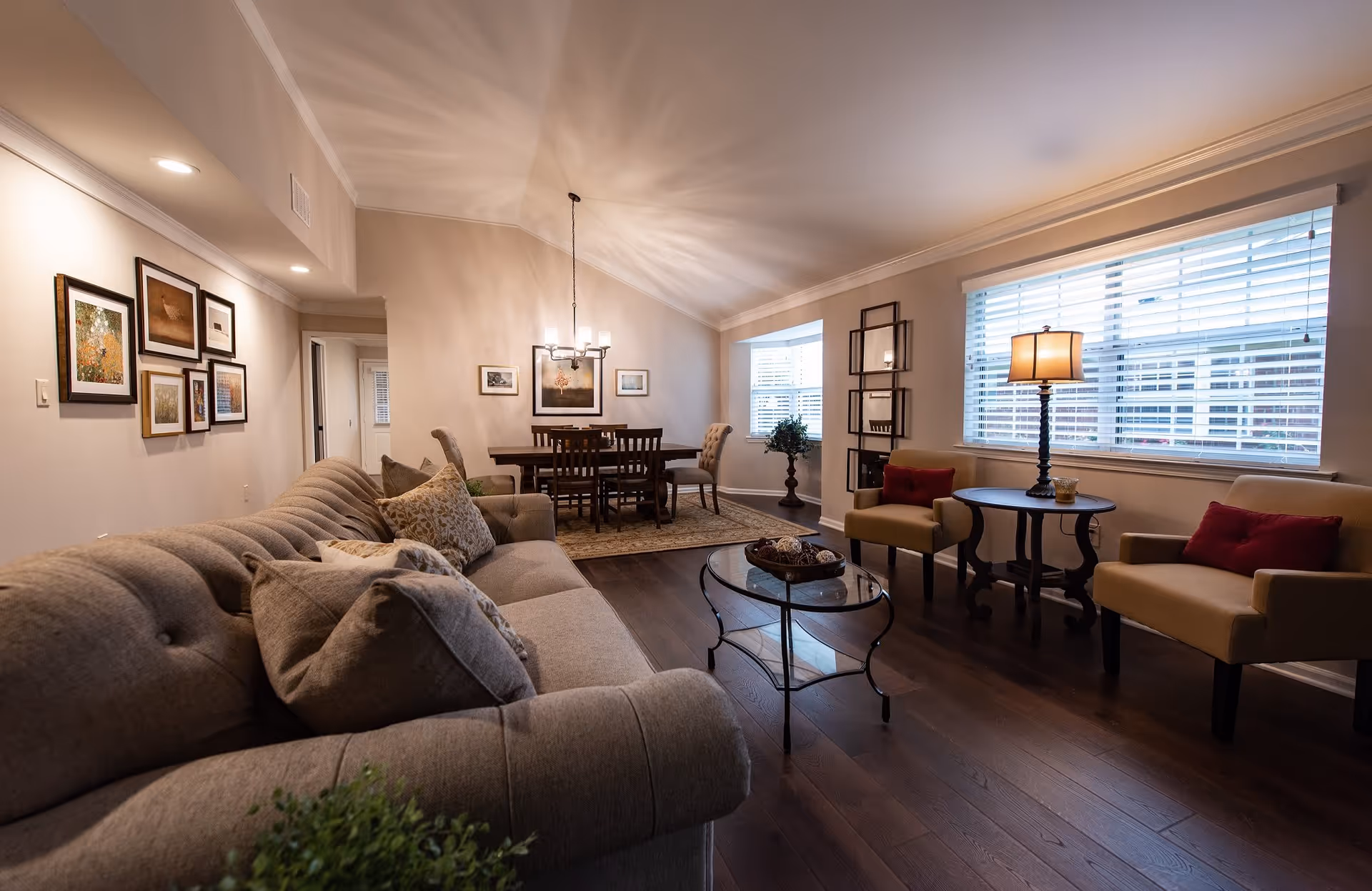 A cozy living room and dining area with a beige tufted sofa adorned with patterned pillows, two beige armchairs with red cushions, a glass coffee table, and a round side table with a lamp. The dining area features a wooden table with six chairs and a chandelier above. The room has wooden flooring, framed artwork on the walls, and large windows with white blinds allowing natural light to enter.