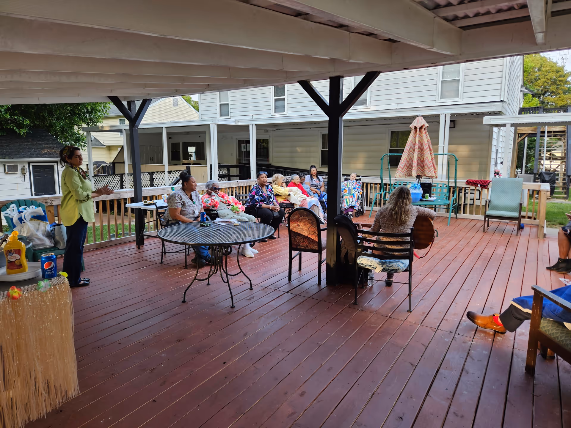 People seated on a covered wooden deck outside a residential building, with one person playing guitar.