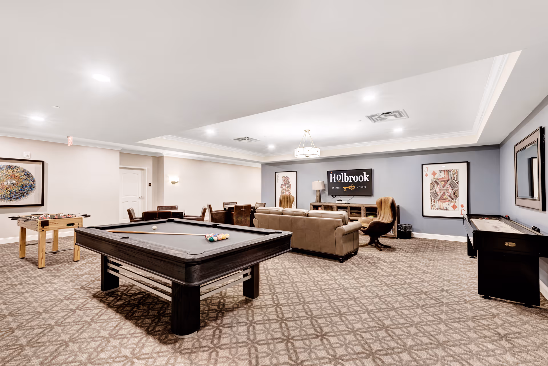 A spacious recreational room with a pool table in the center, a foosball table to the left, and a shuffleboard table to the right. There is a seating area with a beige sofa and two brown chairs facing a wall-mounted TV displaying the Holbrook logo. The room has patterned carpet flooring, framed artwork on the walls, and recessed ceiling lights.