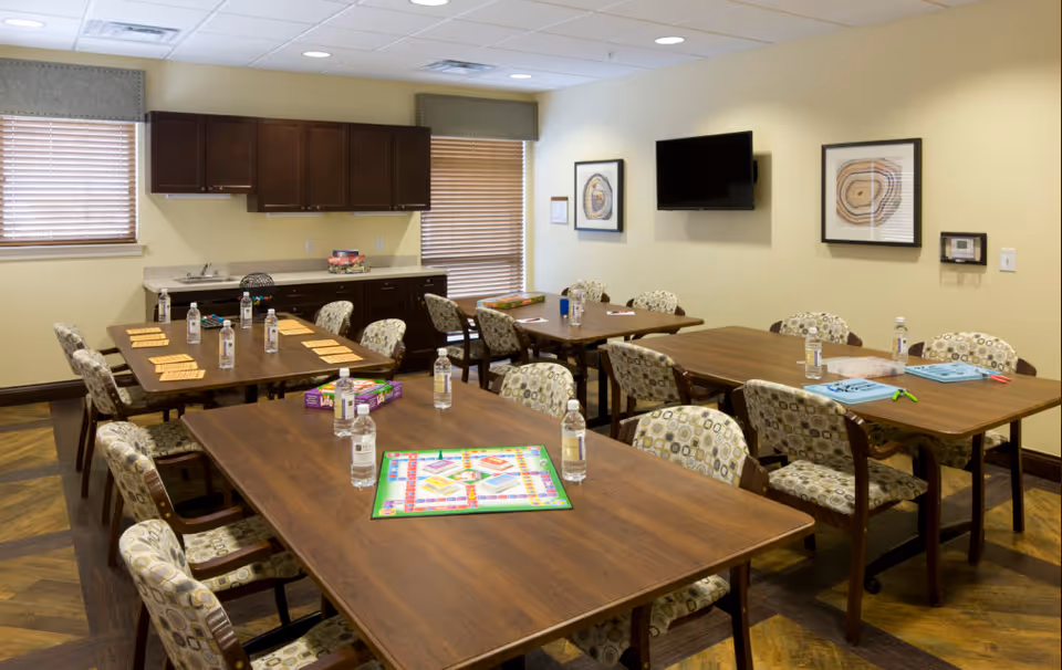 A bright activity/dining room with several wooden tables and patterned chairs, bottled water and board games on the tables, a wall-mounted TV and cabinets.