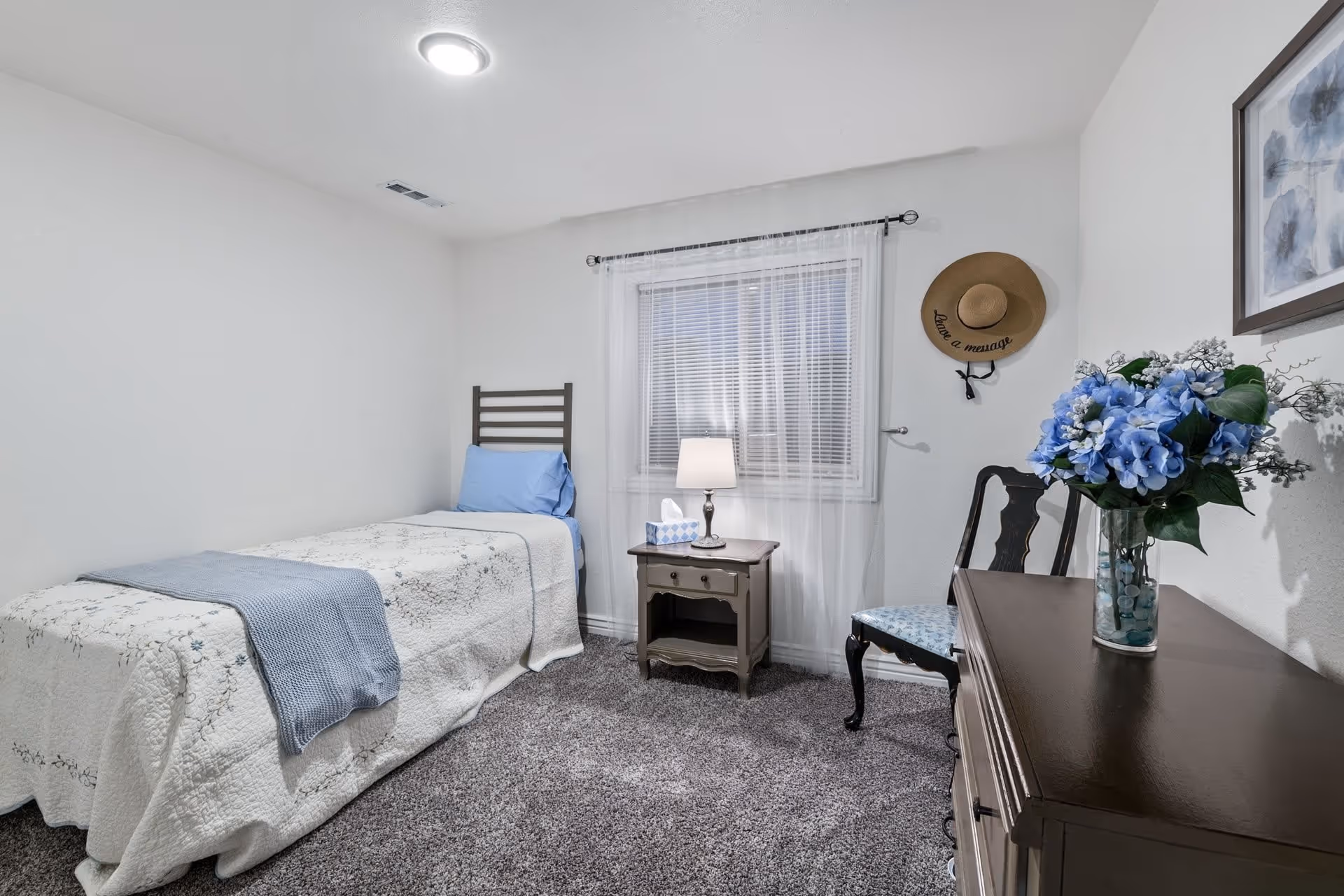 A small, cozy bedroom with a single bed covered in a white quilt with floral patterns and a light blue throw blanket. There is a blue pillow on the bed. Next to the bed is a wooden nightstand with a lamp and a tissue box. A window with sheer white curtains is behind the nightstand. On the right side, there is a wooden chair with a blue patterned cushion and a dresser with a vase of blue flowers. A decorative hat with the words 'Leave a message' hangs on the wall above the chair. The room has light-colored walls and carpeted floor.