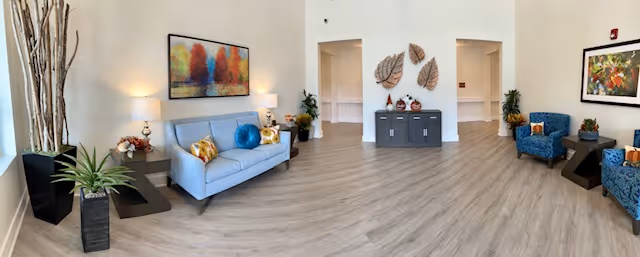 A spacious and well-lit living room area with light wood flooring, a light blue sofa with colorful pillows, two blue armchairs with orange pillows, and modern wall art. There are plants in black pots and decorative items on a gray console table against a white wall.