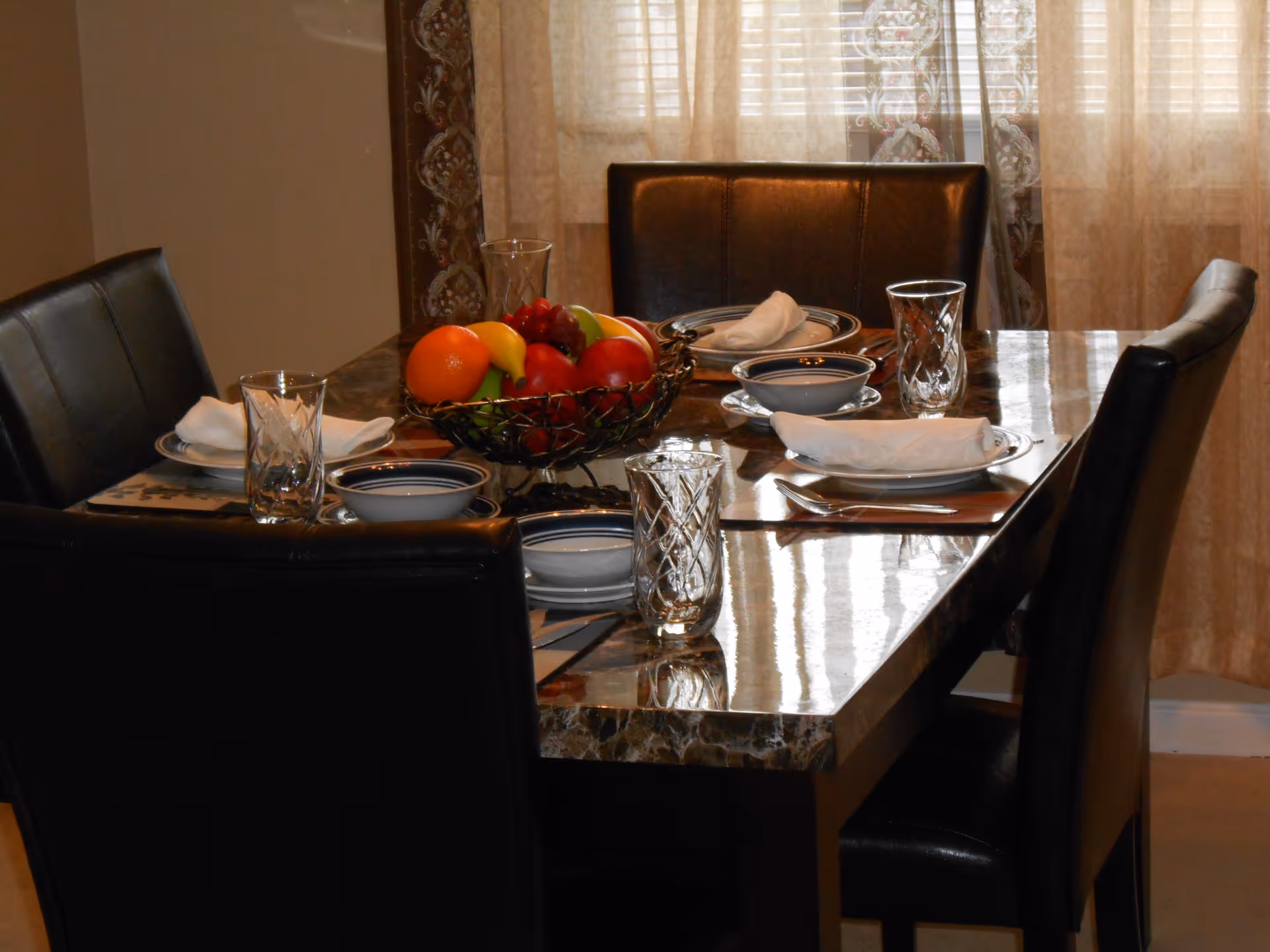 A dining table set for four with black leather chairs, white plates with napkins, bowls, glass tumblers, and a centerpiece bowl filled with assorted fruits including oranges, bananas, apples, and grapes. Sheer curtains with a decorative pattern hang in the background.