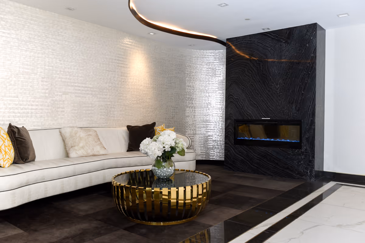 A modern living room with a curved white sofa adorned with various pillows, a round gold and glass coffee table with a vase of white flowers, and a black marble fireplace with a built-in electric fire feature. The walls have a textured light-colored finish and the floor is a combination of dark and light tiles.