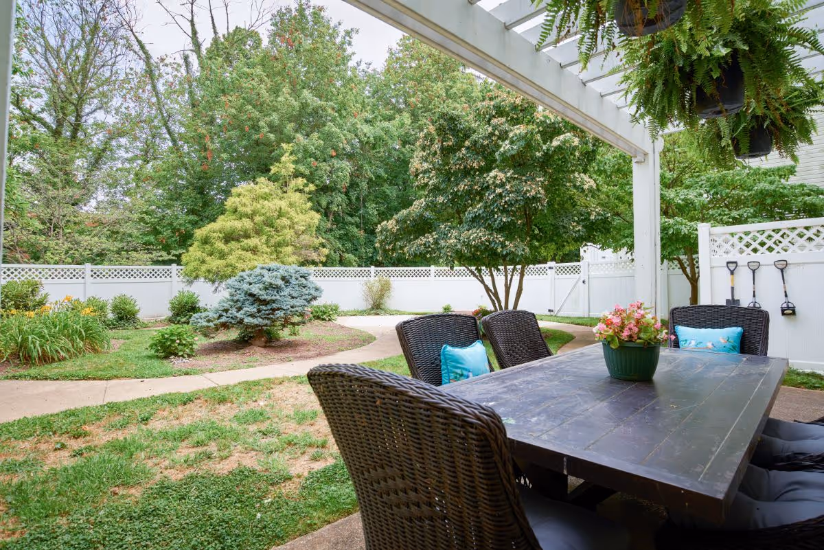 Outdoor patio area with a dark wooden table surrounded by wicker chairs with blue cushions. A green potted plant with pink flowers is on the table. The patio is covered with a white pergola, and hanging plants are visible. In the background, there is a white fence, a paved walkway, and various trees and shrubs in a garden setting.