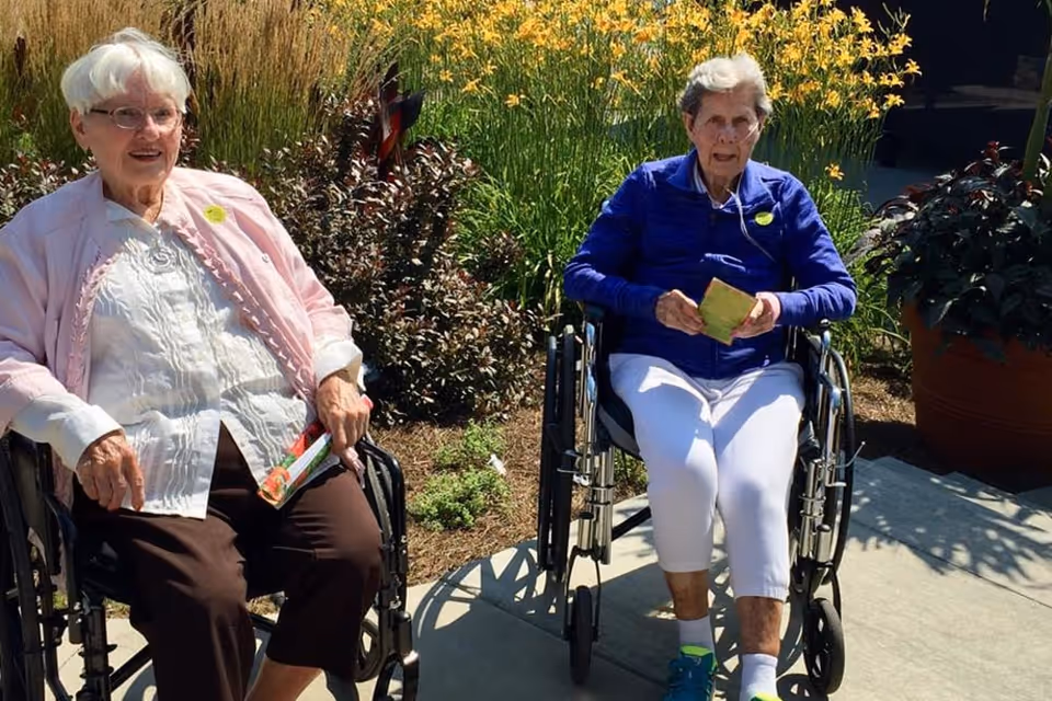 Two elderly women sitting outdoors in wheelchairs on a sunny day, surrounded by plants and flowers. One woman is wearing a light pink jacket and dark pants, while the other is dressed in a blue jacket and white pants.