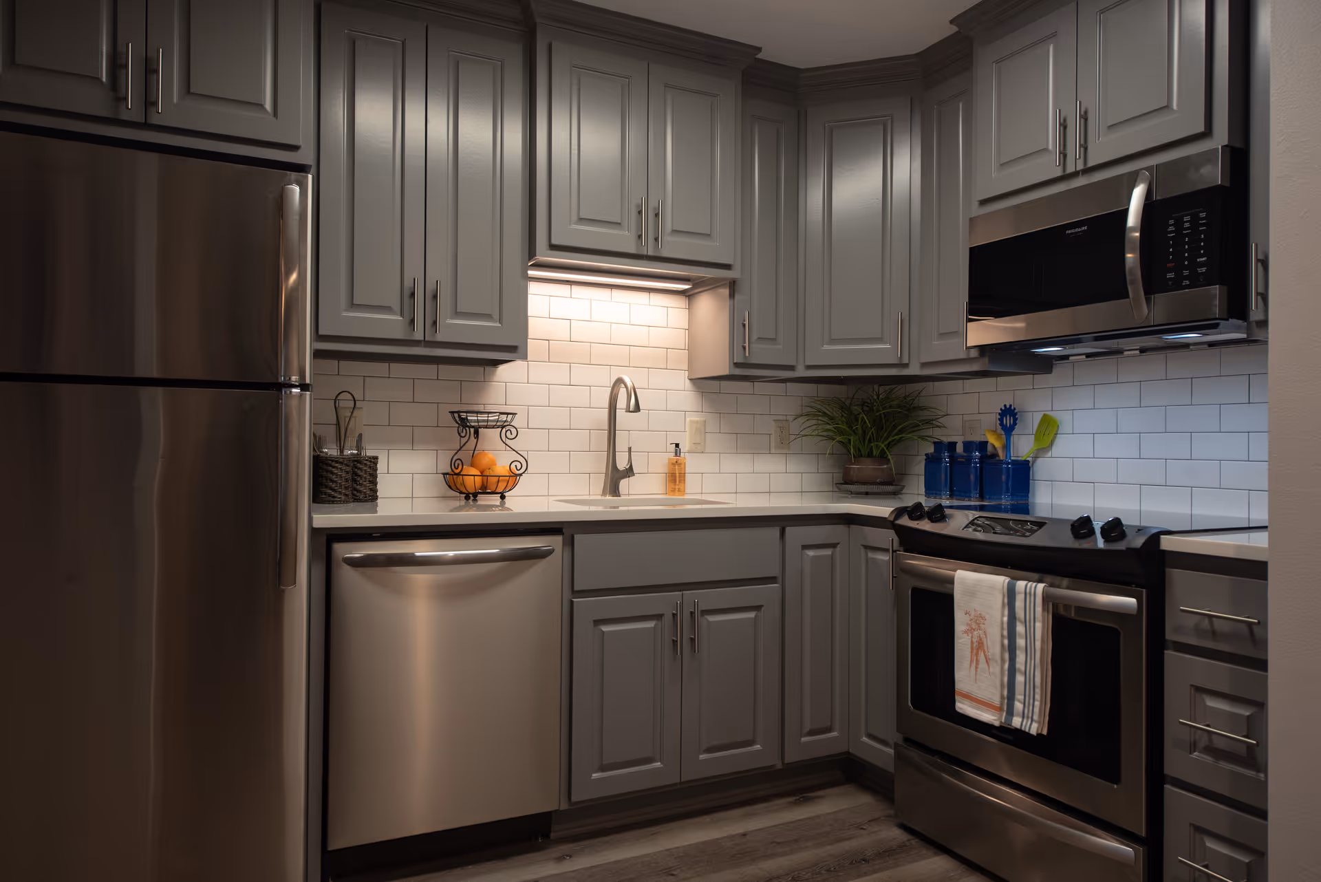 Modern kitchen with gray cabinets, stainless steel refrigerator, dishwasher, oven, and microwave. The backsplash features white subway tiles, and there is a small plant and kitchen utensils on the countertop.