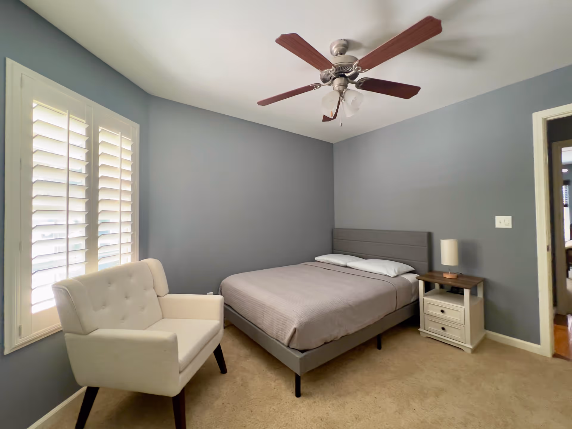 A bedroom with gray walls and beige carpet featuring a bed with gray bedding and two pillows, a white cushioned armchair near a window with white shutters, a wooden nightstand with a lamp, and a ceiling fan with wooden blades.
