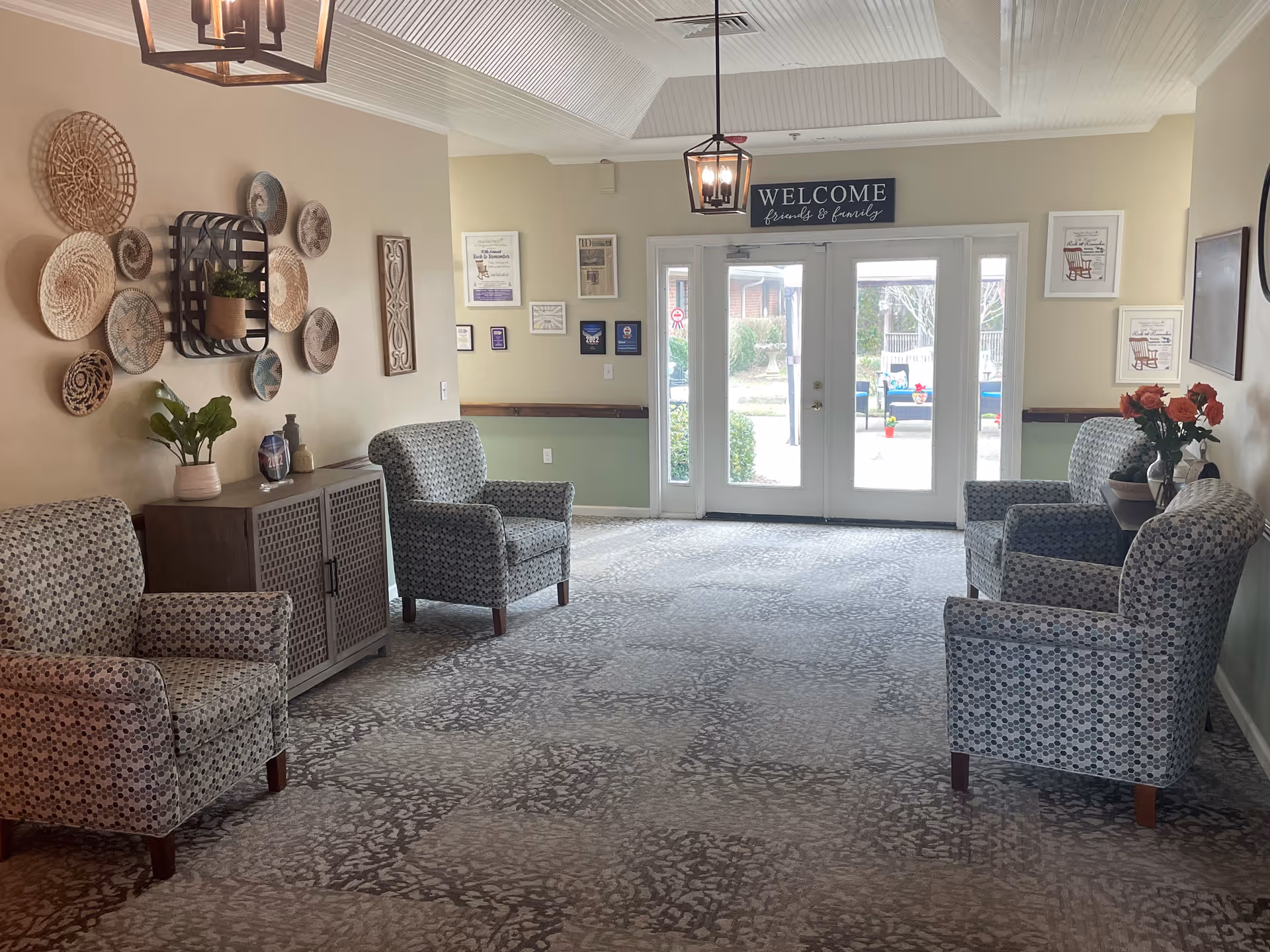 A welcoming seating area with four patterned armchairs arranged along the walls, a cabinet with decorative items and plants, wall art including woven baskets and framed pictures, and double glass doors with a 'WELCOME friends & family' sign above them leading outside.