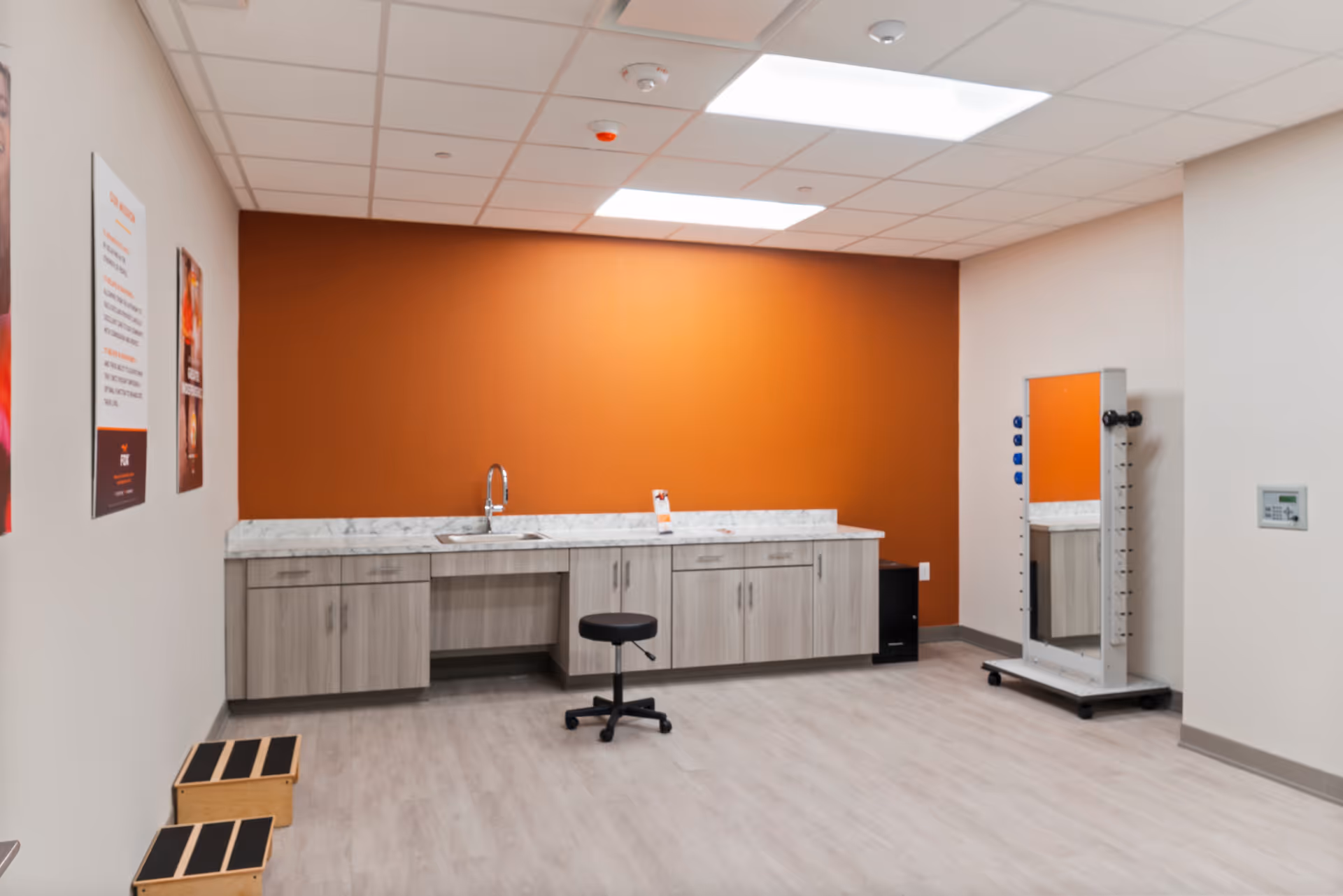 A clean, modern room with light wood flooring and a bright orange accent wall. There is a long countertop with a sink and multiple cabinets underneath. A black swivel stool is positioned in front of the counter. On the right side of the room, there is a tall mirror with blue and black knobs on its frame, mounted on a wheeled base. Two small wooden step stools with black anti-slip surfaces are visible on the left side. The ceiling has recessed lighting and a smoke detector.