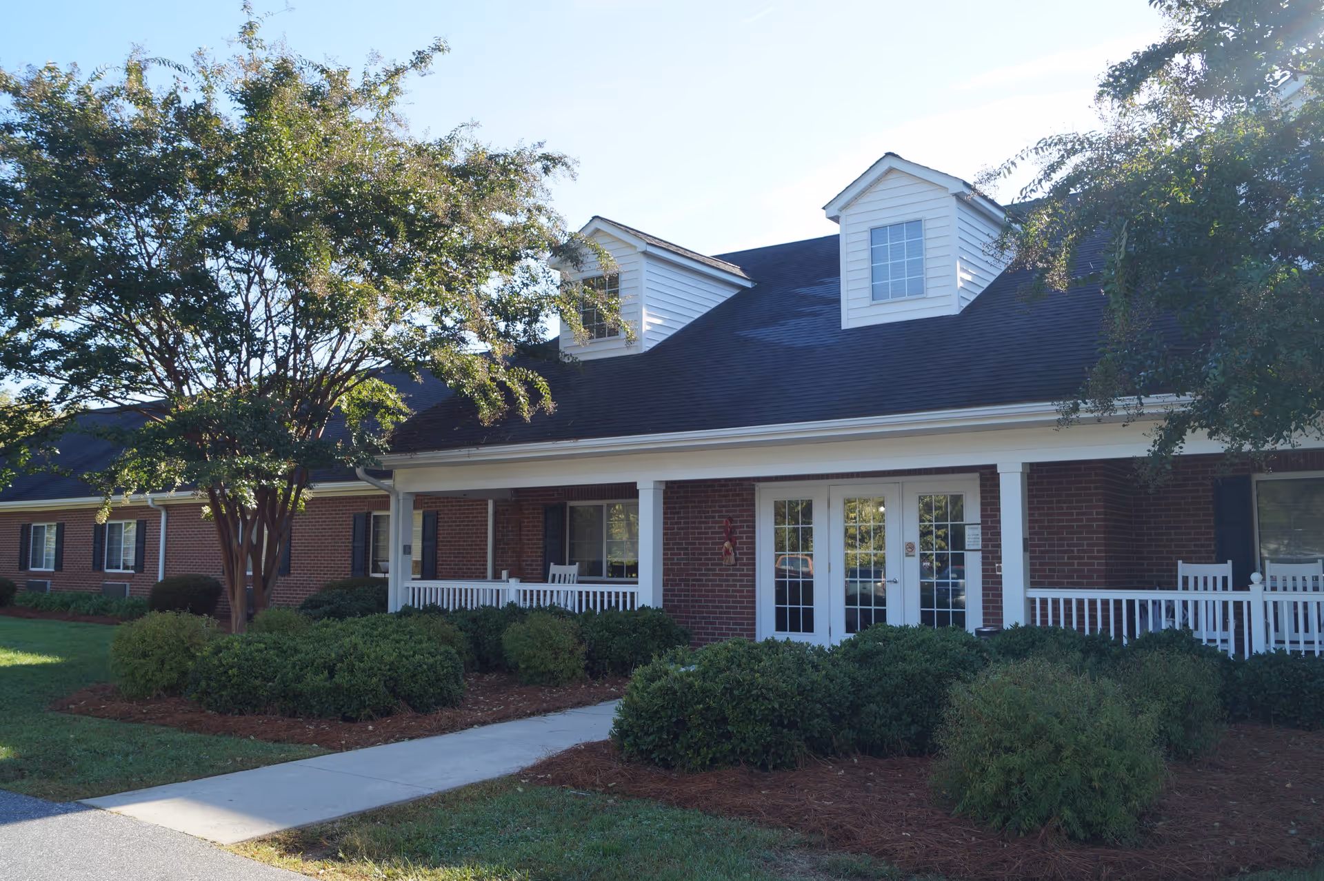 Exterior view of Taylorsville House, a single-story brick building with white-framed windows and doors, a black roof with dormer windows, a covered porch with white railings and rocking chairs, surrounded by green bushes and trees under a clear sky.