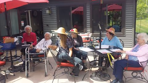 Five elderly women sitting outdoors on a patio at a senior living facility. They are seated around tables with red umbrellas, enjoying a sunny day. One woman is using a walker, and the others are engaged in conversation or looking around. The background shows the exterior of the building with windows and a door.