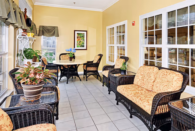 A bright indoor seating area with wicker furniture including cushioned chairs and a loveseat, a round table with chairs, potted plants on tables and shelves, beige walls, tiled floor, and large windows with white frames.