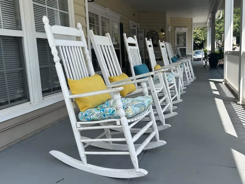 A row of white wooden rocking chairs with colorful cushions lined up on a covered porch outside a building, with windows and a railing visible along the porch.