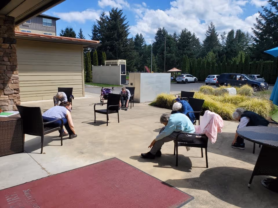 A group of elderly individuals seated on chairs outdoors on a concrete patio, stretching forward towards their feet. The patio is adjacent to a building with beige siding and stone accents. In the background, there are trees, parked cars, and some landscaping with bushes and grass under a partly cloudy sky.