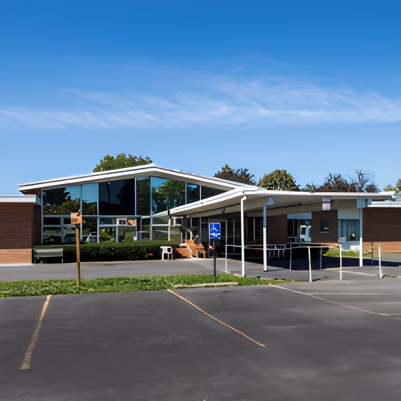 Exterior view of Auburn Rehabilitation and Nursing Center showing a modern building with large reflective windows, a covered entrance with a ramp, and a parking lot in the foreground under a clear blue sky.