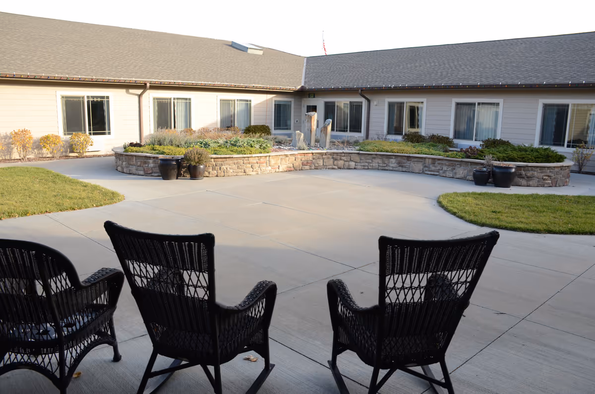 Three wicker chairs on a covered patio facing a paved courtyard with a raised stone planter and single-story building windows.