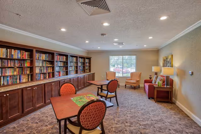 Community library/lounge with bookshelves along one wall, tables and chairs, and armchairs by a window.