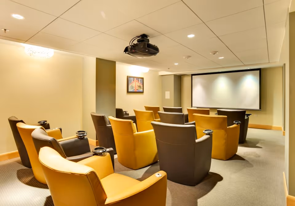 A small theater room with two rows of comfortable armchairs in yellow and brown facing a large projection screen. A ceiling-mounted projector is visible above the chairs, and a framed picture hangs on the wall. The room has soft lighting and a carpeted floor.