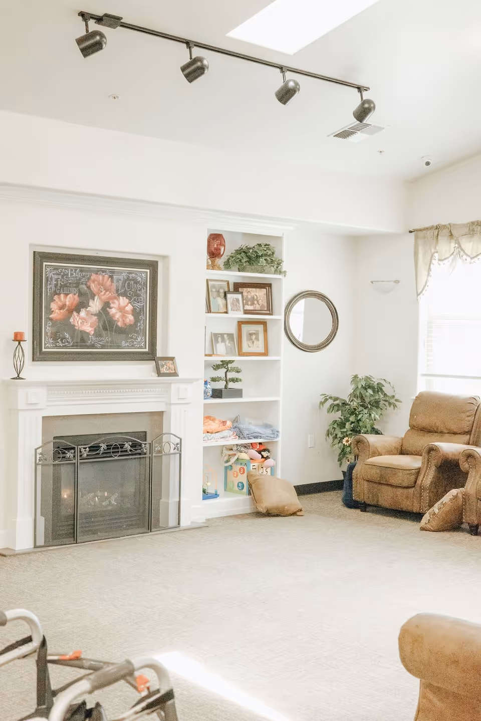 A bright and cozy living room with a white fireplace featuring a floral painting above it. Next to the fireplace is a built-in white shelving unit with framed photos, plants, and blankets. There is a round mirror on the wall and a large window with sheer curtains letting in natural light. Two brown upholstered armchairs are positioned near a green potted plant. A walker is partially visible in the foreground.