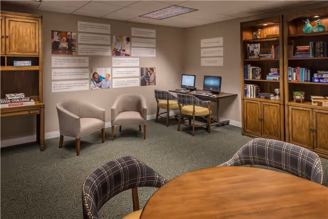 A cozy senior living common area with a round wooden table and plaid upholstered chairs in the foreground. Two beige armchairs are placed against the wall, near a desk with two computers and chairs. Wooden bookshelves filled with books and decorative items line the right side of the room. The walls have framed quotes and pictures, and the carpet has a subtle patterned design.