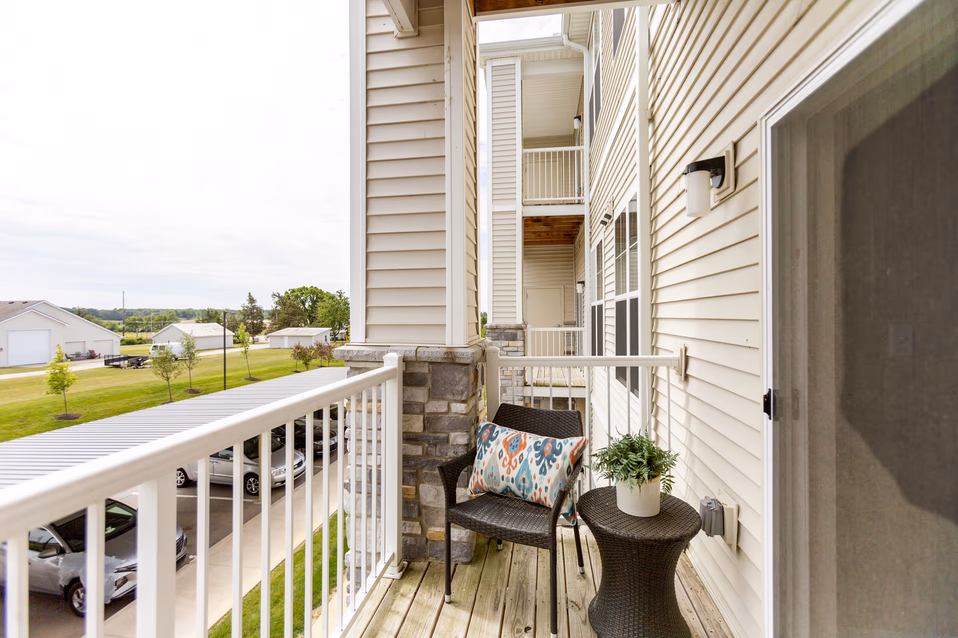 A small balcony with a wicker chair featuring a colorful patterned cushion and a matching round wicker table with a potted plant. The balcony has white railings and overlooks a parking area with cars and a grassy area with trees and buildings in the distance.