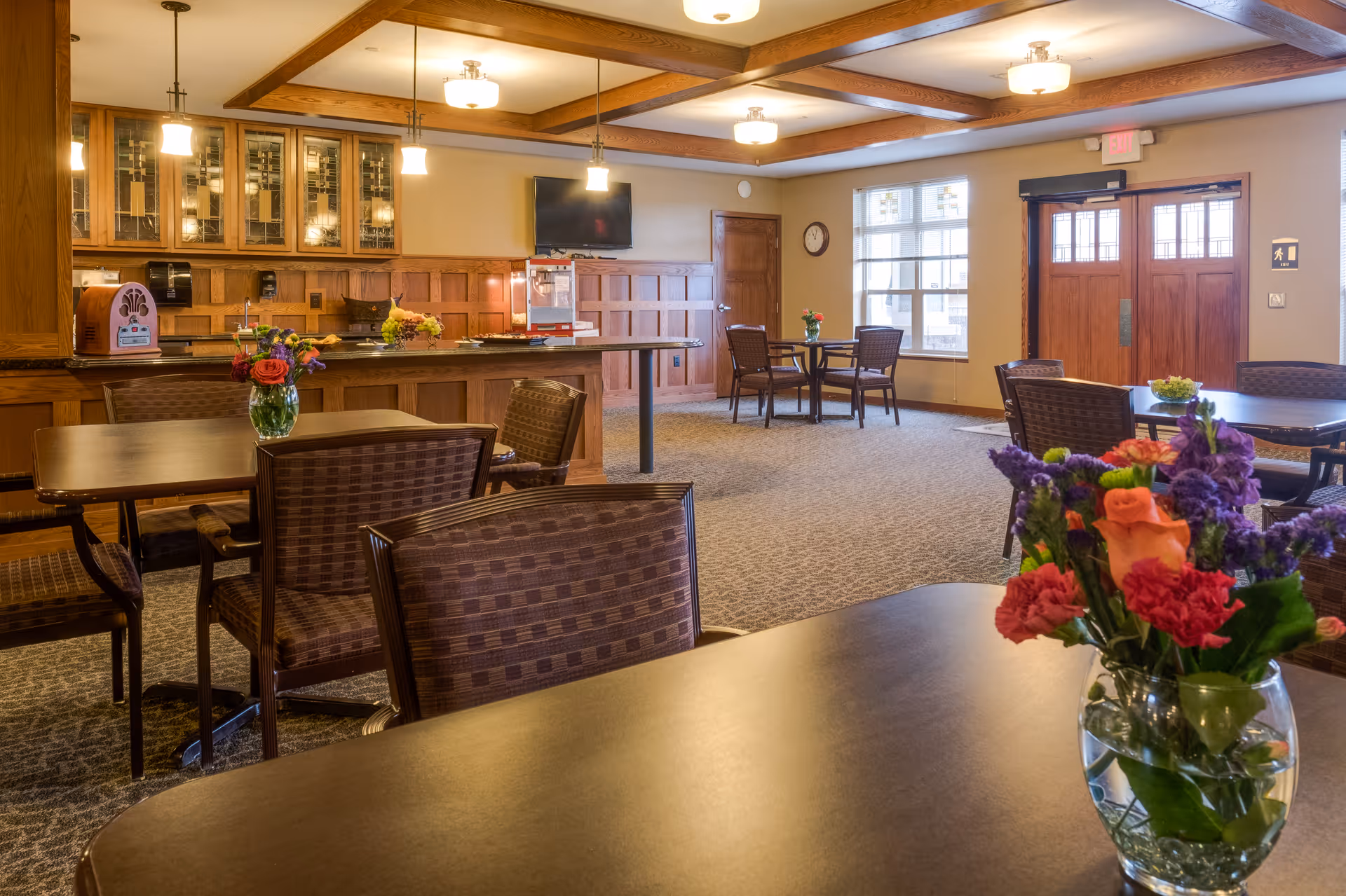 A warm and inviting dining area in a senior living facility with multiple tables and chairs. Each table has a vase with colorful flowers. The room features wooden paneling, a counter with a popcorn machine, a wall-mounted TV, and large windows letting in natural light. The ceiling has wooden beams and hanging lights.