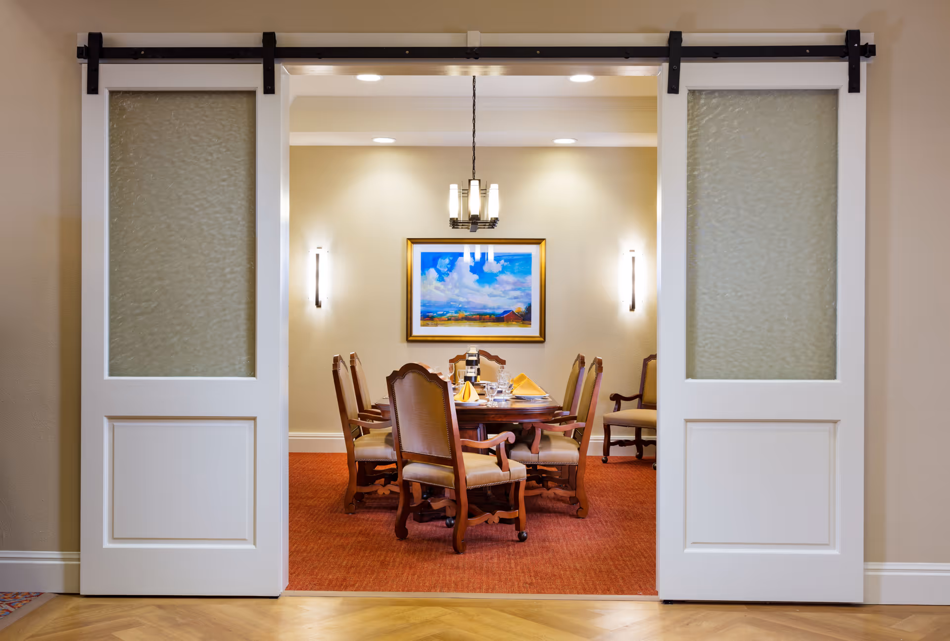 View through white sliding doors into a dining room with a wooden table set for six, upholstered chairs, a chandelier overhead, wall sconces, and a framed painting of a landscape on the far wall.