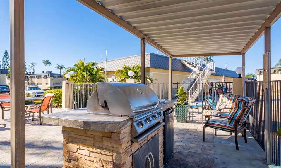Outdoor covered patio area with a stainless steel barbecue grill, a striped cushioned chair, and a view of a swimming pool and stairs in the background under a clear blue sky.
