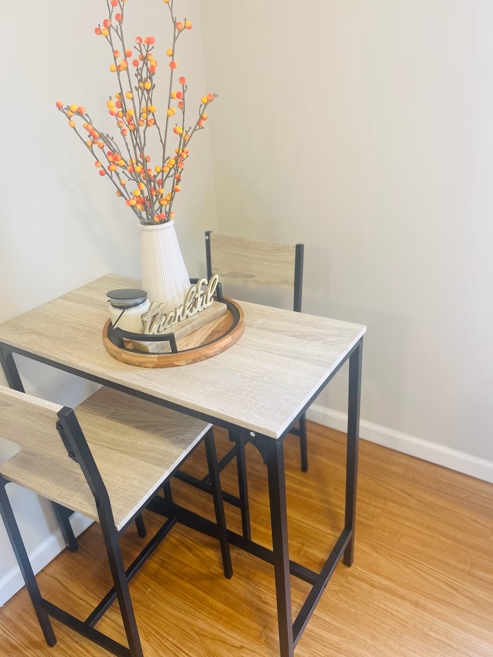 A small wooden dining table with black metal legs and two matching chairs on a wooden floor. On the table is a white vase with decorative branches bearing orange and yellow berries, a candle, and a wooden tray with a decorative sign that says 'thankful'. The background is a plain light-colored wall.