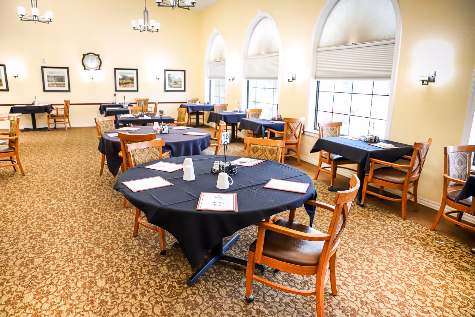 Bright dining room with round and square tables covered in black tablecloths, wooden chairs, and arched windows along one wall.