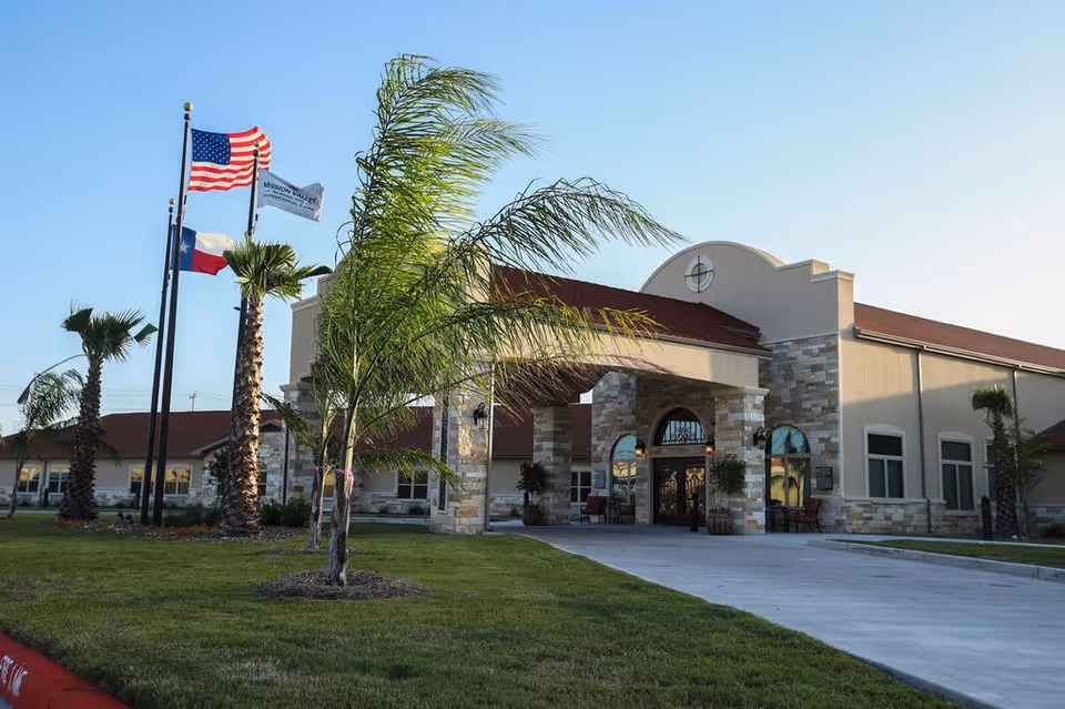 Front entrance of a nursing and transitional care building with flags, palm trees, and a driveway.