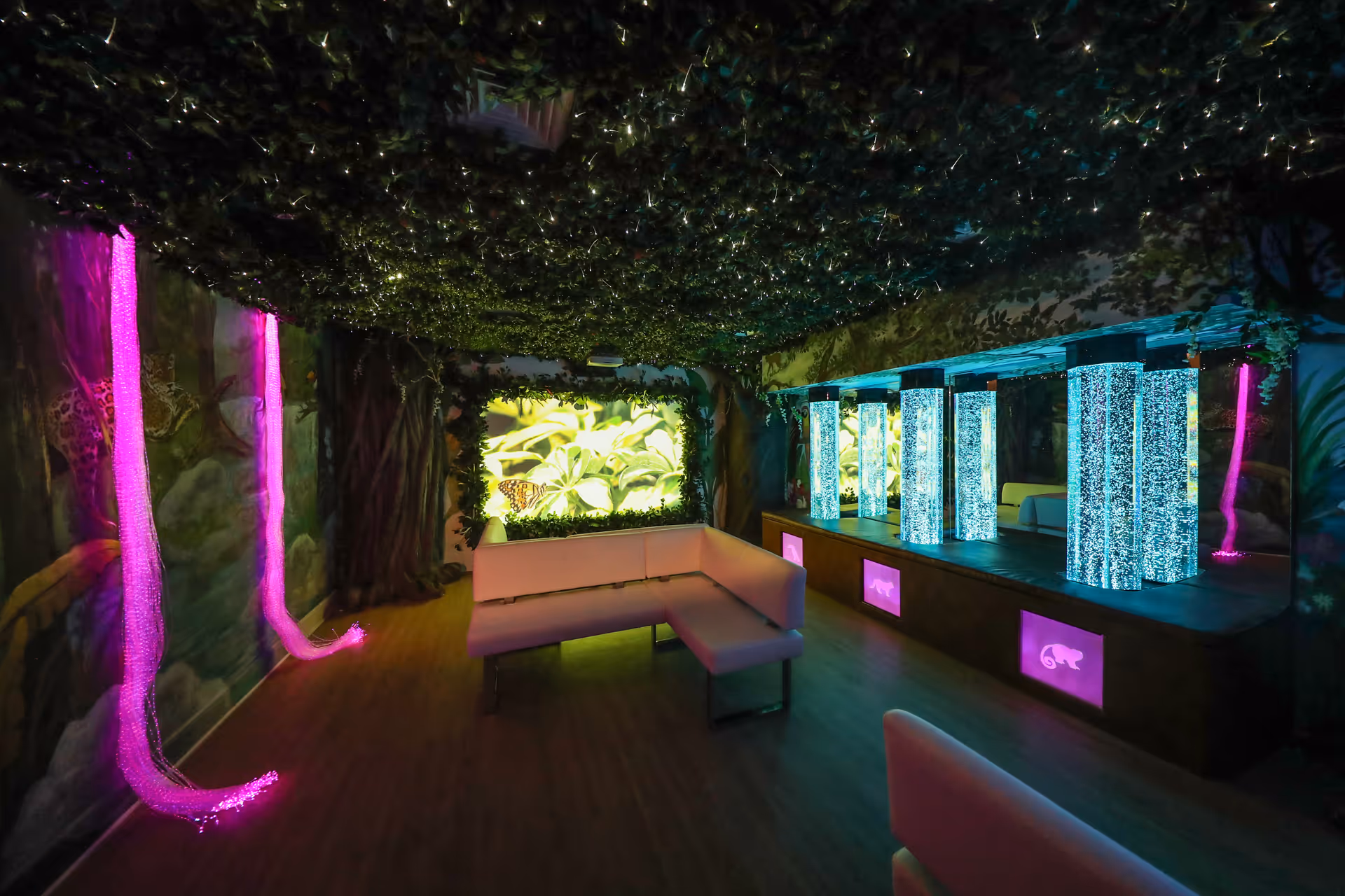 A sensory room with a jungle theme featuring a ceiling covered in artificial greenery, walls painted with forest and animal murals, glowing bubble tubes emitting blue light, and fiber optic lights hanging in pink. There is an L-shaped white cushioned bench in the center of the room.
