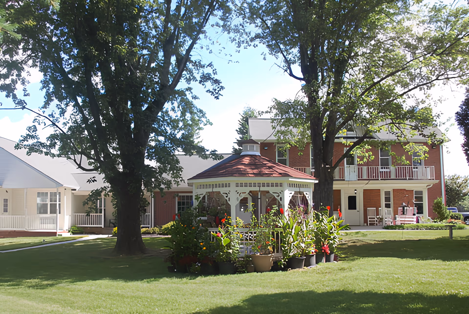 A sunny outdoor garden area at Homestead-Sun Valley Assisted featuring a white gazebo surrounded by potted plants and flowers. Large trees provide shade over a well-maintained green lawn. In the background, there is a two-story brick building with white trim and a porch area with chairs.