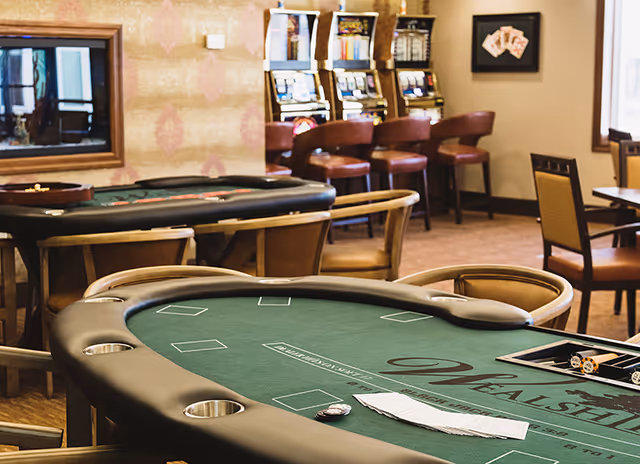 Interior room with a poker table in the foreground, several chairs around it, and slot machines with bar stools in the background. The room has warm lighting and framed artwork on the walls.