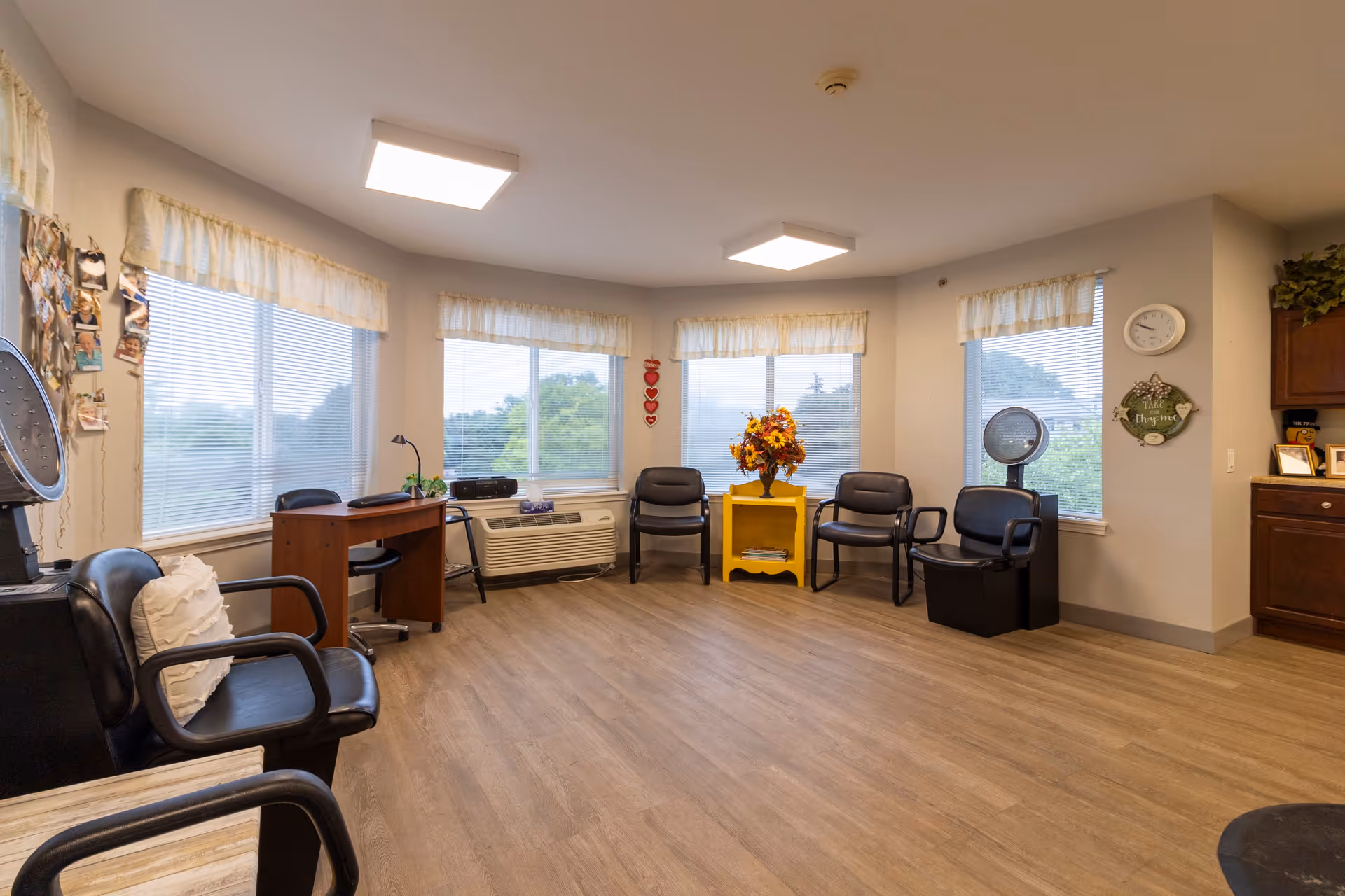 A bright room with large windows covered by light curtains, featuring several black chairs arranged around the perimeter. There is a small wooden desk with a chair, a yellow side table with a flower arrangement, a wall clock, and a hair dryer station. The floor is wood, and the walls are light-colored.