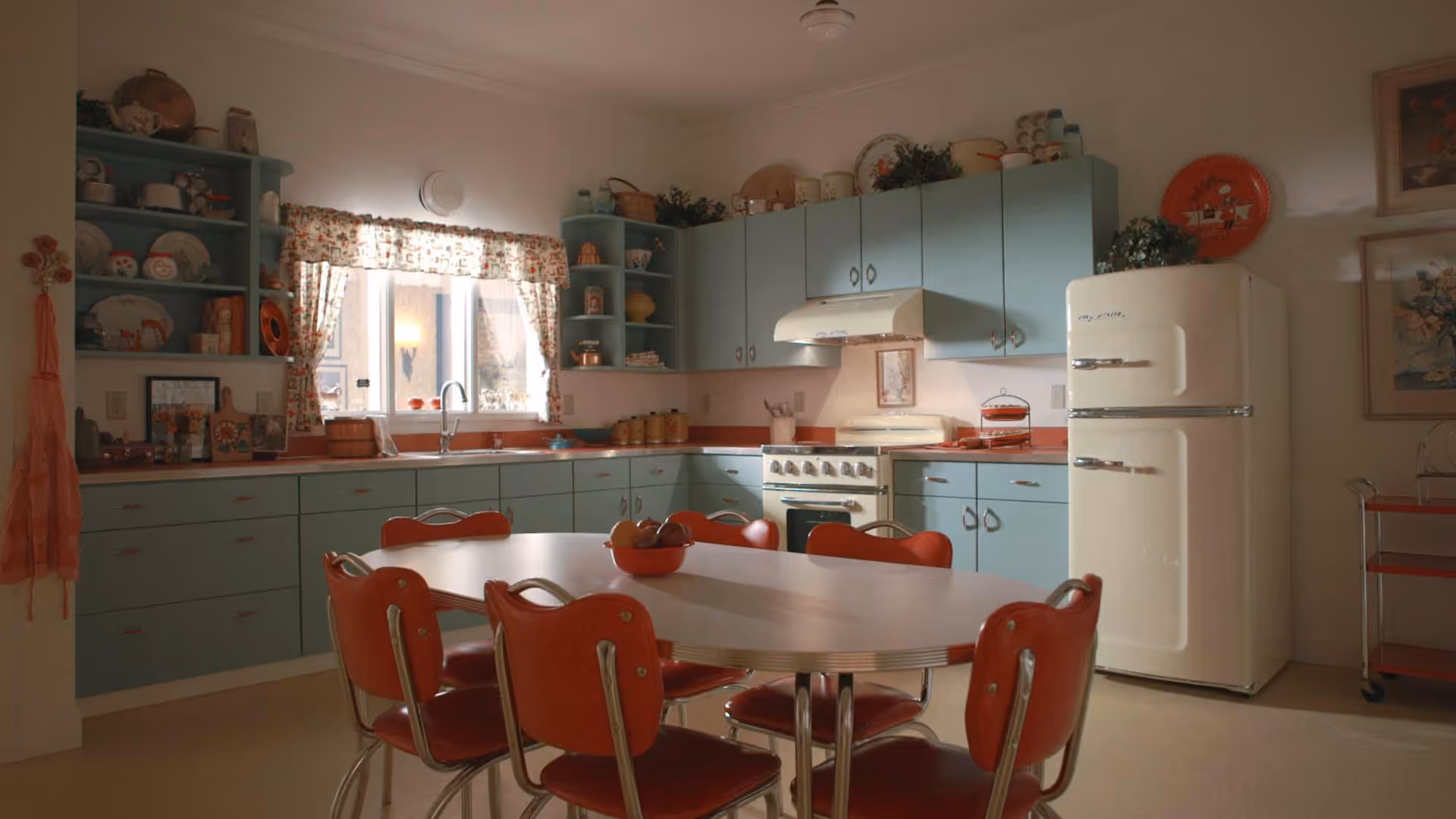 A vintage-style kitchen with light blue cabinets, a white retro refrigerator, and a white stove with a vent hood. There is a window with floral curtains above the sink, and a dining table with six red chairs in the center of the room. Various kitchen items and decorations are displayed on the shelves and countertops.