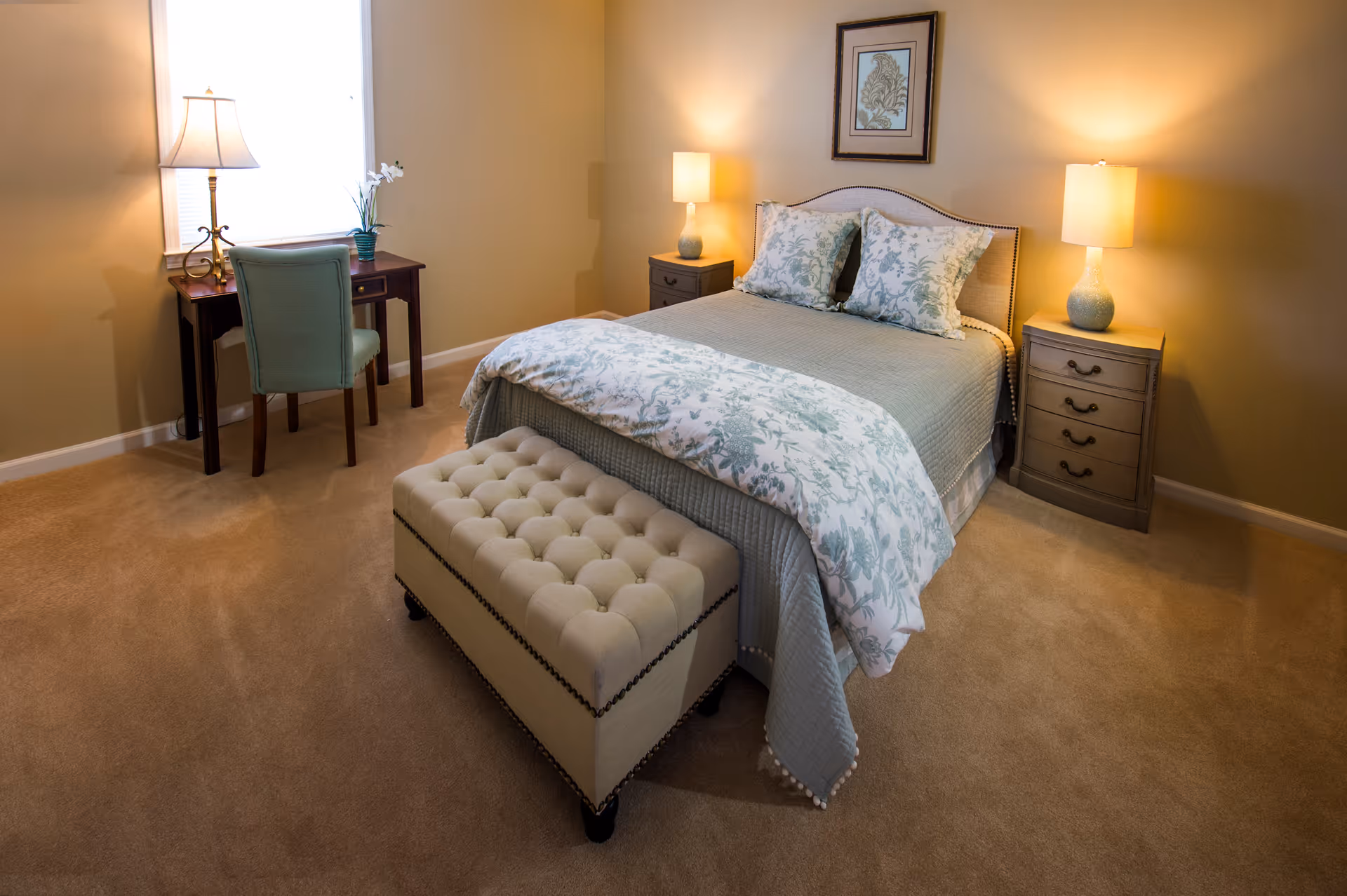 Neatly arranged bedroom with bed, nightstands, lamps, a tufted ottoman, and a desk.