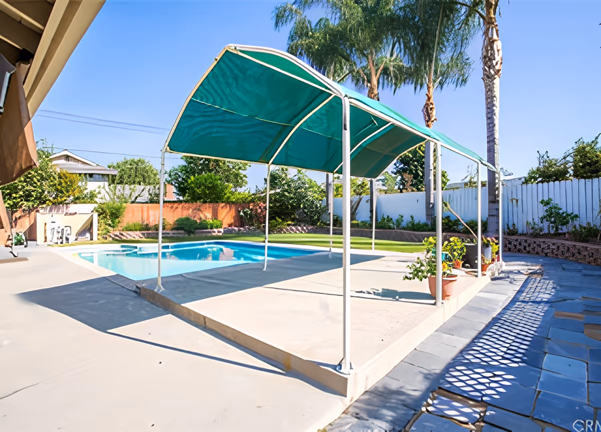 Outdoor area with a swimming pool, a green canopy shade structure, potted plants, palm trees, and a white fence surrounding the yard under a clear blue sky.