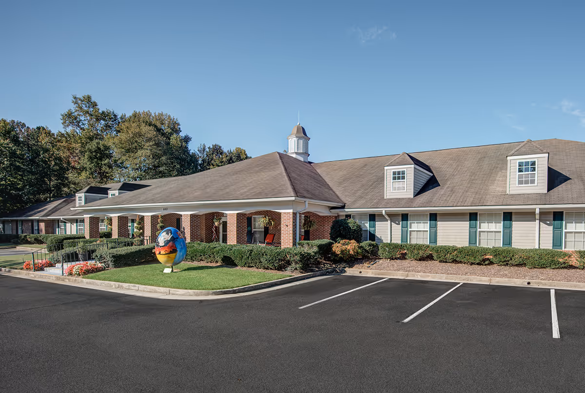 Exterior view of a senior living facility building with a brick and siding facade, a cupola on the roof, green shutters on the windows, and a small landscaped area with bushes and a colorful egg-shaped sculpture near the entrance. The sky is clear and blue, and there is an empty parking lot in front.
