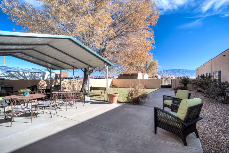 Outdoor courtyard with a covered patio, metal dining tables and chairs, wicker lounge chairs with green cushions, a large tree and distant mountain views.