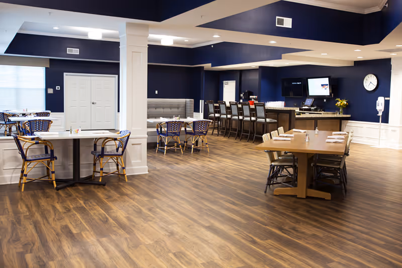 Spacious dining area with multiple tables and chairs, a bar counter, and wood flooring under navy blue walls.