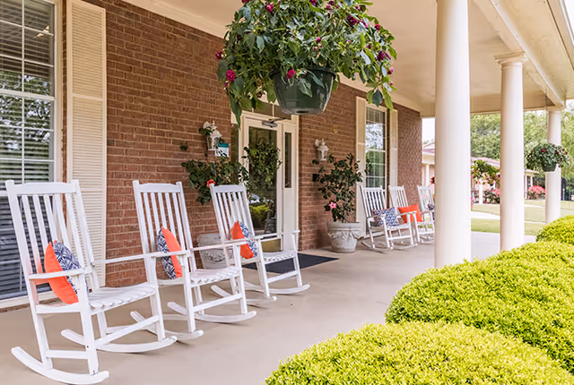 A covered porch area with white rocking chairs lined up against a brick wall. Each chair has a decorative pillow, and there are hanging flower pots and potted plants along the porch. Green bushes are visible in the foreground.